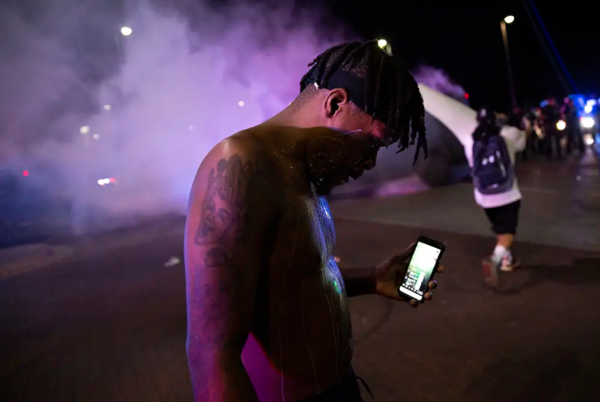 A protester covered in milk livestreams himself while Dallas Police tactical officers fire rounds of tear gas during a march in memory of  George Floyd on May 29, 2019.